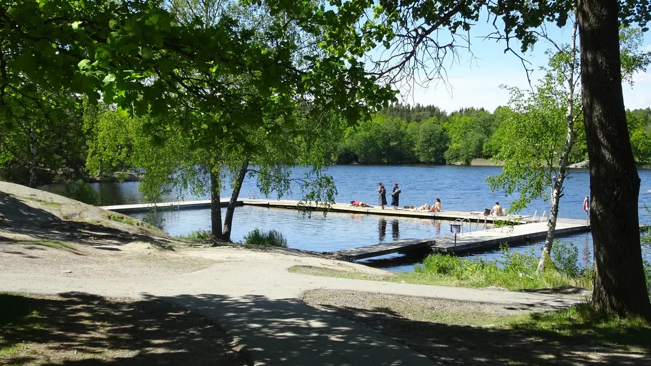 Badbrygga vid sjö med badande, träd och grön skog i bakgrunden